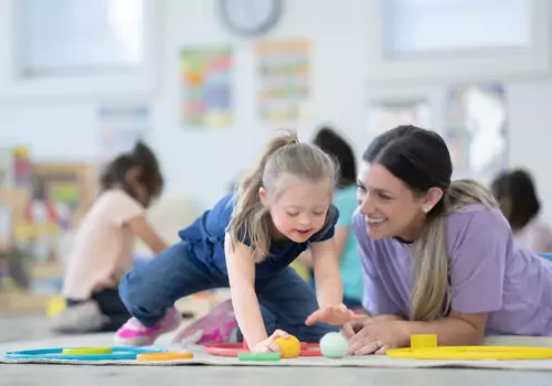A smiling Preschool Teacher from Morton IL lays on the floor with a child to work on a project