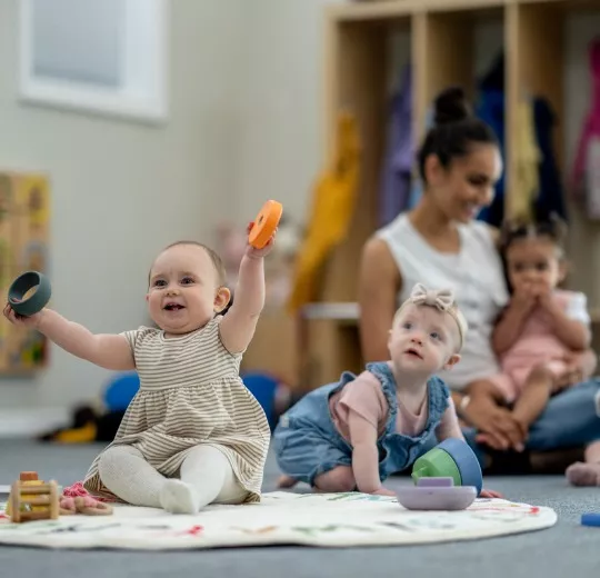Two babies sit on the floor playing as part of the PALS infant program while their teacher and other PALS babies are in the background
