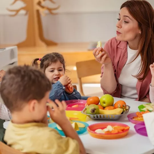 A teacher smiles warmly as she interacts with children enjoying a healthy snack at a table during PALS’ Two’s and Three’s Program