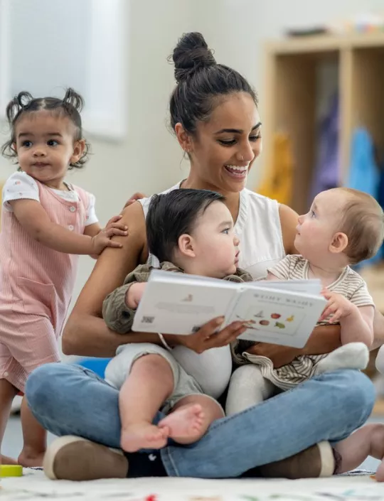 A teacher sits with a large group of babies as she read them a book in their classroom. Our Programs & Curriculum provide experiences with caring teachers.