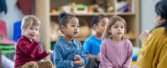 A small group of children in PALS’ Pre-K Program sit on the floor in front of their teacher as she leads them in a song.