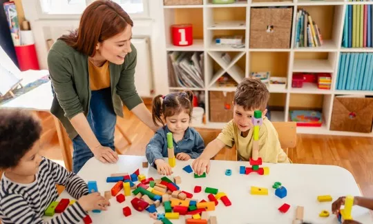 A smiling teacher interacts with a group of young children playing with colorful blocks in a bright, cheerful classroom in PALS’ Two’s and Three’s Program.